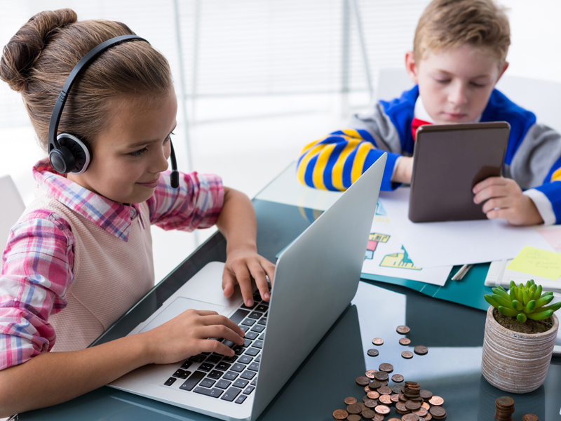 Auf dem Bild befinden sich zwei Kinder. Ein Kind sitzt vor dem Laptop und hat Kopferhörer auf, das zweite Kind hält ein Tablet in der Hand.